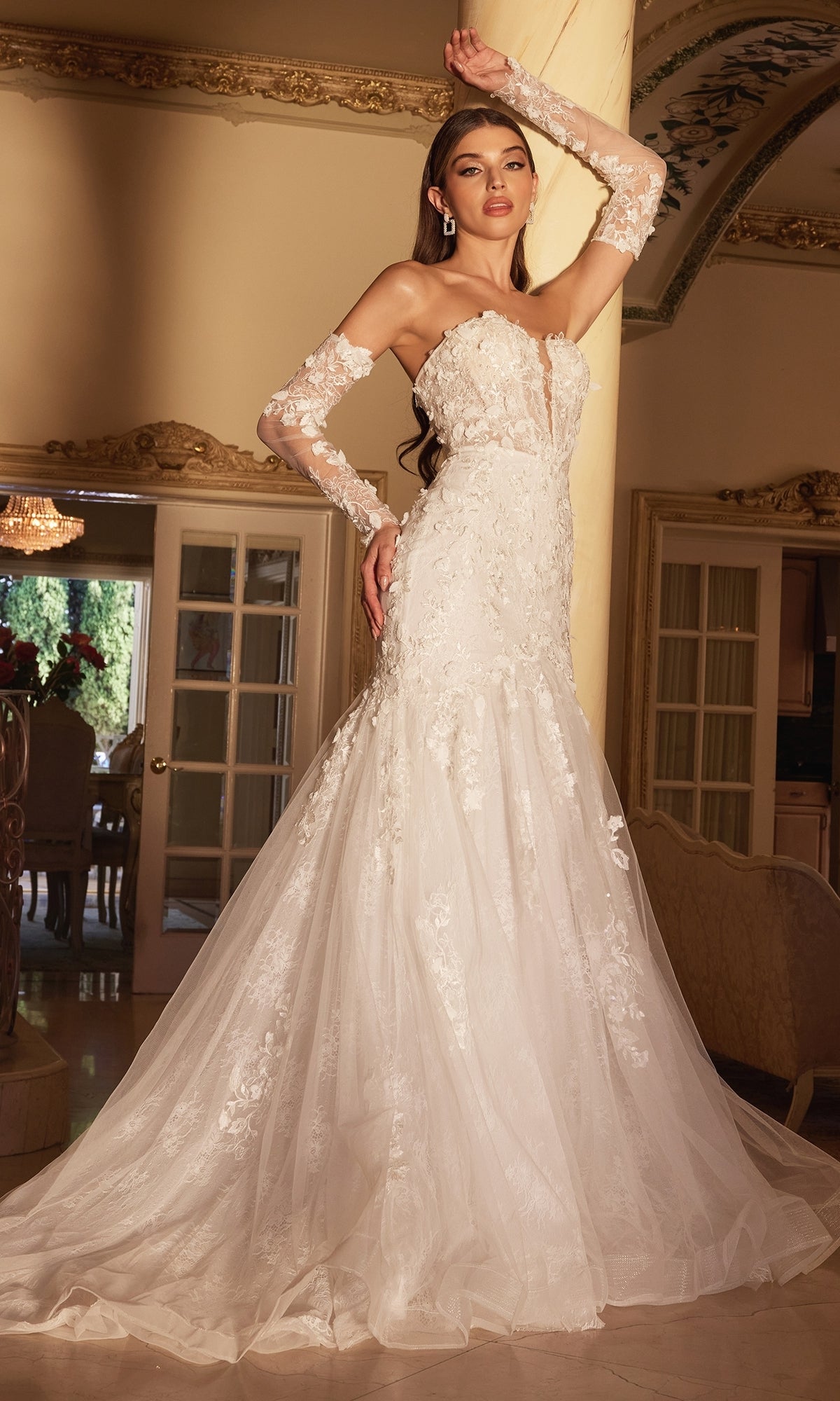 A woman poses indoors with one arm raised, wearing the Long White Strapless Bridal Gown CD977W featuring a fitted bodice, embroidered floral designs, and elegant sheer sleeves.